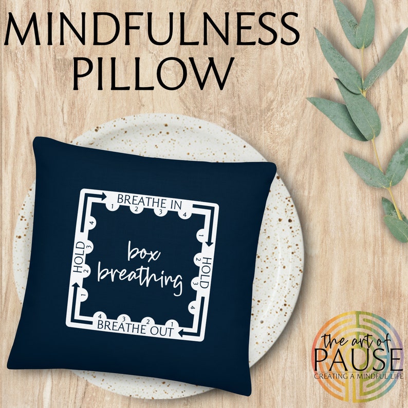 May include: A navy blue pillow with white text and a diagram for box breathing. The text reads "BREATHE IN", "HOLD", "box breathing", "BREATHE OUT", and "HOLD". The pillow is on a white plate with a speckled pattern. The text "the art of PAUSE CREATING A MINDFUL LIFE" is visible in the lower right corner of the image.