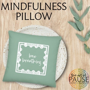 May include: A green pillow with white text that says "Breathe In", "Hold", "Box Breathing", "Hold", and "Breathe Out". The pillow is on a white plate with brown speckles. The text is in a box with arrows showing the direction of the breath.