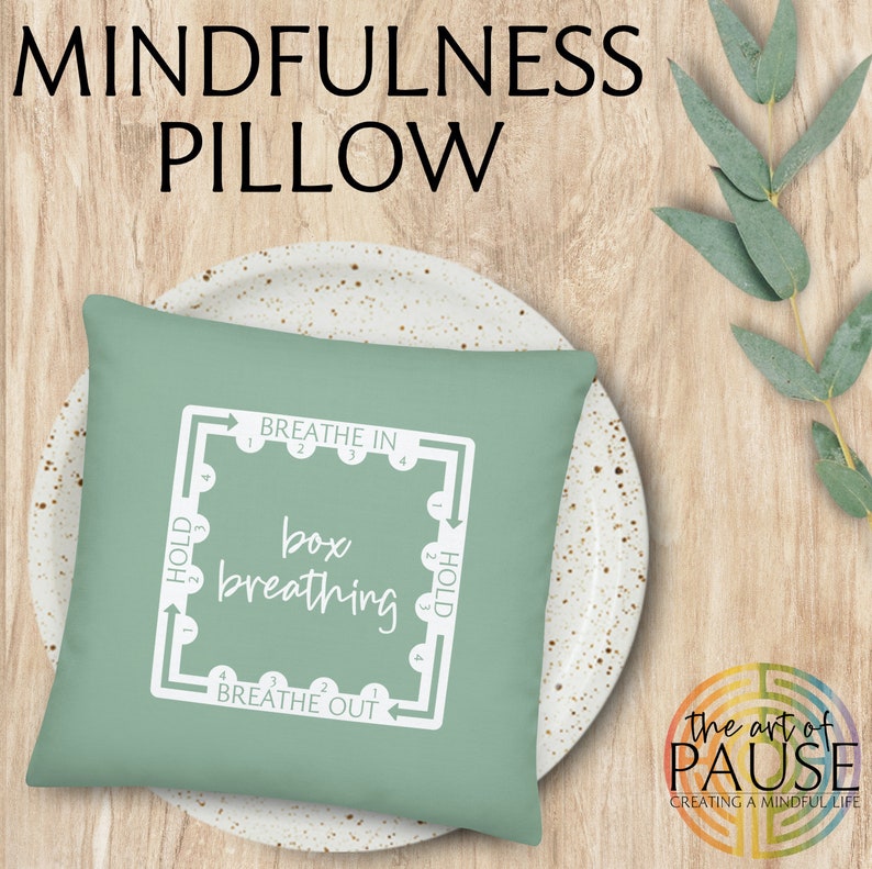 May include: A green pillow with white text that says "Breathe In", "Hold", "Box Breathing", "Hold", and "Breathe Out". The pillow is on a white plate with brown speckles. The text is in a box with arrows showing the direction of the breath.