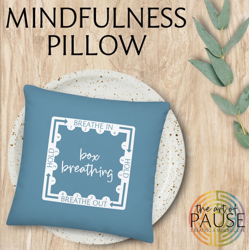 May include: A blue square pillow with white text that reads "BREATHE IN", "box breathing", "BREATHE OUT", and "HOLD". The text is arranged in a square with arrows pointing in a clockwise direction. The pillow is on a white plate with a speckled pattern.
