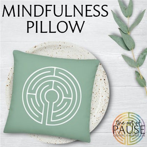 May include: A green square pillow with a white circular labyrinth design. The pillow is on a white plate with brown speckles. The text "the art of PAUSE creating a mindful life" is in the lower right corner of the image.