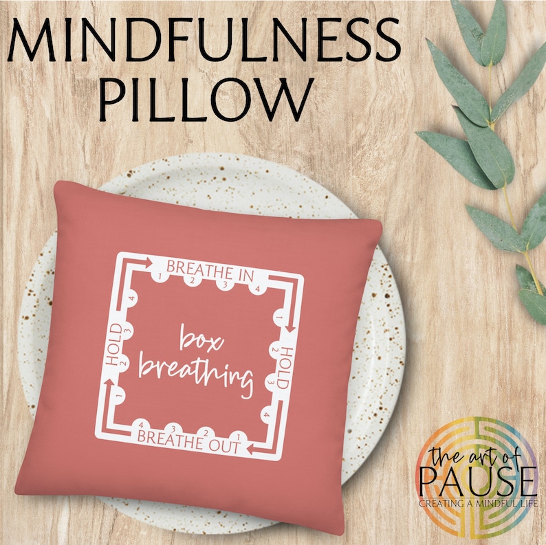 May include: A coral-colored square pillow with white text and a diagram for box breathing. The text reads "Breathe In", "Hold", "Breathe Out", "Hold", and "box breathing". The pillow is on a white plate with brown speckles. The text "the art of PAUSE creating a mindful life" is in the lower right corner of the image.
