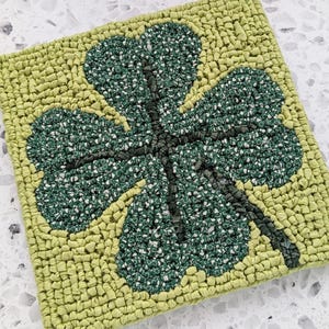May include: A green four-leaf clover rug made of looped fabric. The clover is surrounded by a light green background.