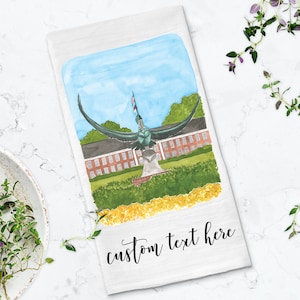 May include: A white tea towel with a watercolor illustration of a statue of a bird with outstretched wings. The statue is in front of a brick building with a flagpole and an American flag. The text "custom text here" is written in black cursive at the bottom of the towel.