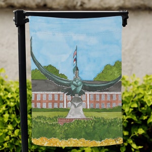 May include: A watercolor painting of a garden flag featuring a stylized eagle with outstretched wings, standing on a pedestal with a flagpole and American flag. The eagle is in front of a brick building with windows and a green lawn.