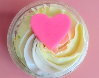 Könnte beinhalten: Ein klares Glasgefäß gefüllt mit einem Wirbel aus weißem und gelbem geschlagenem Körperbutter, gekrönt mit einer rosa herzförmigen Seife.