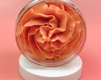May include: A clear, circular plastic jar filled with a thick, peach-coloured whipped cream substance. The product is piped into a swirling, rose-like pattern that rises above the rim of the container, which rests on a solid white circular lid.