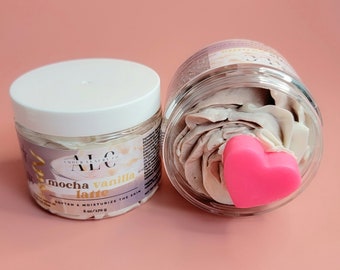 May include: Two clear jars of body butter with white lids. One jar is upright, the other is tilted, revealing the contents. The body butter is a light brown color and topped with a pink heart. Text on the jar reads "mocha vanilla latte" and "soften & moisturize the skin".
