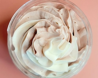 Può includere: Un contenitore di plastica trasparente riempito con un vortice di panna montata in tre colori: bianco, marrone chiaro e marrone più scuro. Il contenitore è su una superficie rosa. La panna montata ha un aspetto liscio e strutturato.