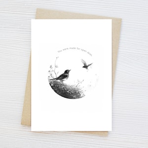 Może przedstawiać: Kartka okolicznościowa w czerni i bieli z ilustracją dwóch ptaków. Jeden siedzi na gnieździe, drugi leci. Tekst „You were made for open skies” znajduje się nad nimi. Kartka leży na brązowej kopercie.