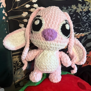 May include: A handmade, pink crochet plush toy with large black eyes, long ears, and a purple nose. The plush has a white belly and is sitting on a red pillow. The toy is a character from the Lilo & Stitch franchise.