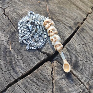 Può includere: Una collana con catena d'argento con un ciondolo in osso con una pila di teschi intagliati. Il ciondolo ha la forma di un cucchiaio.