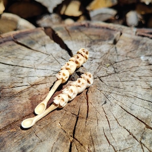 Può includere: Due cucchiai di legno intagliati con una serie di motivi a forma di teschio. I cucchiai sono appoggiati su una superficie di legno.