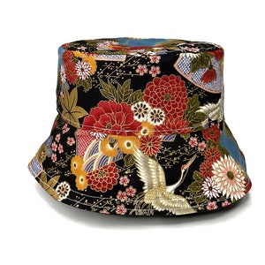 Op de afbeelding: Een zwarte bucket hat met een kleurrijke bloemen- en kraanvogelprint. De print bevat rode, gele en witte bloemen, gouden accenten en een witte kraanvogel.