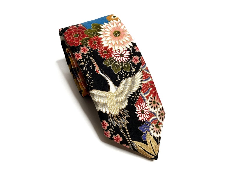 May include: A necktie featuring a black background with a detailed pattern of white cranes, large pink and red chrysanthemum flowers, and green leaves. The design includes gold-colored accents and small floral motifs, creating a vibrant, multi-colored appearance across the fabric surface of the folded necktie.