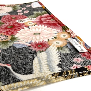 May include: Close-up of a fabric item with a Japanese-inspired floral and crane design. The fabric features a black background with red, pink, and white flowers, and a white crane. The item has a tag that reads "FUJITA SAN".