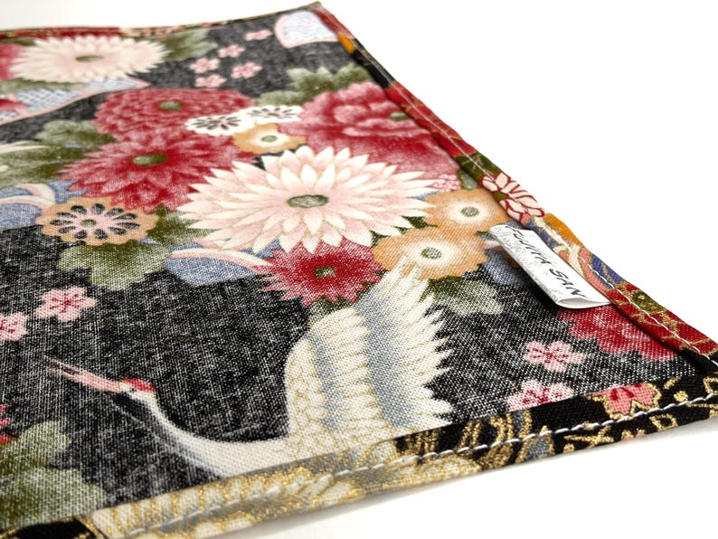May include: Close-up of a fabric item with a Japanese-inspired floral and crane design. The fabric features a black background with red, pink, and white flowers, and a white crane. The item has a tag that reads "FUJITA SAN".