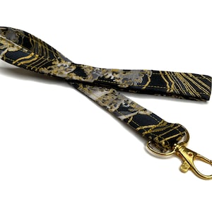 Puede incluir: Un cordón negro y dorado estampado con un cierre dorado. El cordón presenta un diseño repetitivo de elementos florales dorados y grises sobre un fondo negro. El cierre es de estilo giratorio.