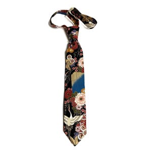 May include: A necktie featuring a black background with a vibrant pattern of white cranes, red and pink chrysanthemums, and gold accents. The fabric displays a detailed floral and bird motif with a blue patch, finished with a pointed tip.