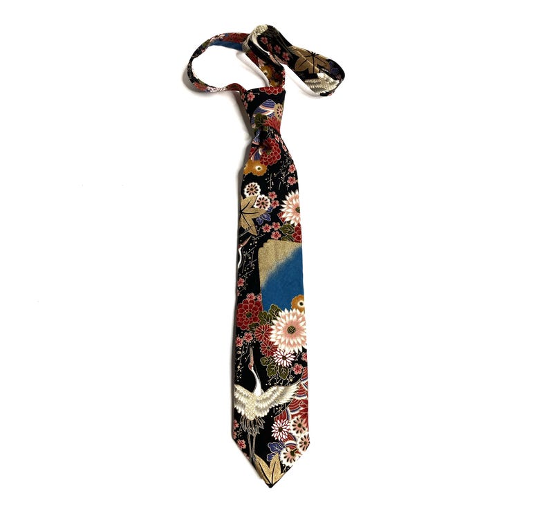 May include: A necktie featuring a black background with a vibrant pattern of white cranes, red and pink chrysanthemums, and gold accents. The fabric displays a detailed floral and bird motif with a blue patch, finished with a pointed tip.
