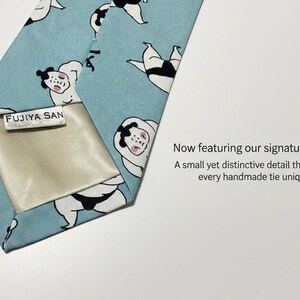 May include: A light blue necktie featuring a repeating pattern of sumo wrestlers in black and white with pink accents. The back of the tie includes a cream-colored fabric lining and a white rectangular label with the text FUJIYA SAN printed in black.