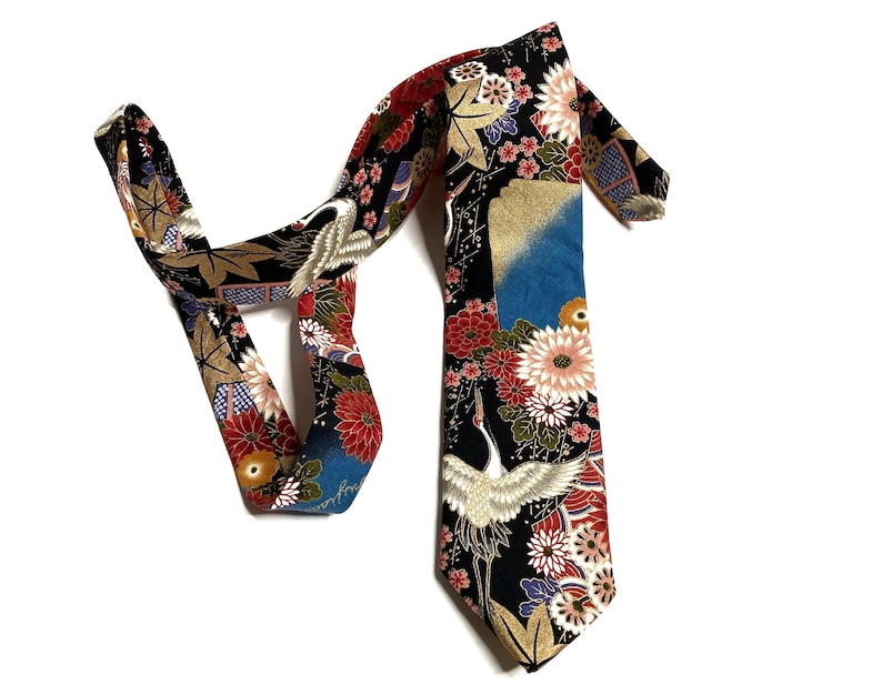 May include: A black necktie featuring a vibrant pattern of white cranes, red and white chrysanthemums, gold maple leaves, and blue geometric accents. The fabric displays a detailed floral and bird motif across the entire length of the tie.