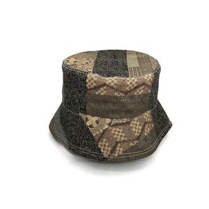Puede incluir: Un sombrero de pescador de patchwork con una mezcla de telas estampadas en tonos negros, marrones y beige. El sombrero tiene una corona redondeada y un ala ligeramente inclinada hacia abajo. Los tejidos incluyen diseños geométricos y texturizados.