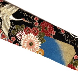 May include: A black fabric featuring a repeating pattern of white cranes with gold accents, interspersed with large red, pink, and white chrysanthemum flowers, green leaves, and blue sections with gold detailing throughout the textile design.