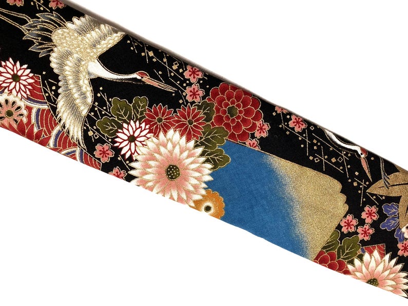 May include: A black fabric featuring a repeating pattern of white cranes with gold accents, interspersed with large red, pink, and white chrysanthemum flowers, green leaves, and blue sections with gold detailing throughout the textile design.