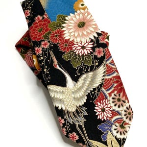 May include: A necktie featuring a black background with a detailed pattern of white cranes, red and pink chrysanthemums, and gold accents. The fabric displays a complex floral and bird motif with blue and green highlights throughout the design.