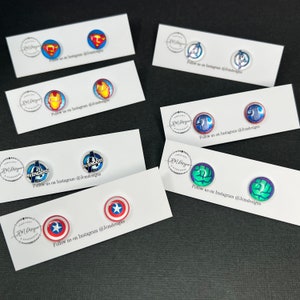 May include: A set of 10 pairs of superhero themed earrings. The earrings are made of resin and feature various Marvel characters, including Captain America, Iron Man, Hulk, and the Avengers logo. The earrings are displayed on white cards with the text "Follow us on Instagram @Jemdesigns".