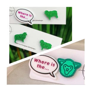 May include: A set of stud earrings featuring green glitter sheep shapes and white speech bubbles with the text Where is the... in pink lettering. The earrings are displayed on white cards with black text and branding details visible on the card surface.