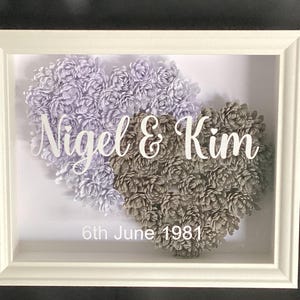 May include: A white framed artwork with two heart-shaped floral arrangements in lavender and grey. The names "Nigel & Kim" and the date "6th June 1981" are displayed in white script. A decorative piece for a wedding or anniversary.