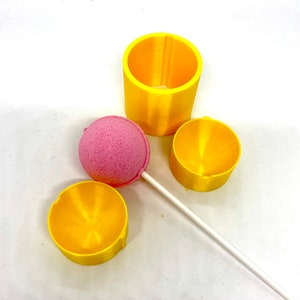 Peut inclure: Trois moules en plastique jaune pour faire des sucettes, un cylindre haut et deux cylindres plus courts, avec une sucette rose sur un bâton blanc.