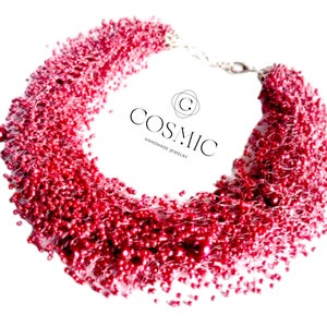 Può includere: Una collana di perline rosse con una chiusura argentata. La collana è realizzata con piccole perline rotonde e ha un design a cascata. Il nome del marchio "COSMIC HANDMADE JEWELRY" è visibile nell'immagine.