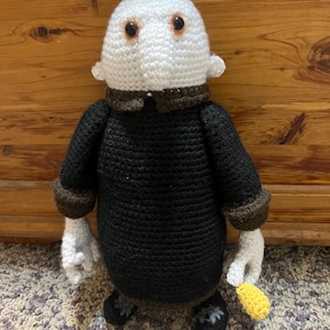 May include: A crocheted plush toy with a white head, black body, and brown accents. The toy has a round head with two large brown eyes and a small nose. The toy is wearing a black jacket with brown trim and is holding a yellow crocheted object in its right hand.