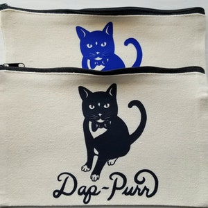 May include: Two canvas zipper pouches with black zippers. One pouch has a blue cat graphic, the other has a black cat graphic. The black cat graphic has the text "Dap-Purr" below it. The pouches are a light beige color.