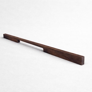 Puede incluir: Un tirador de armario de madera marrón oscuro. El tirador es rectangular con un área de agarre empotrada. La veta de la madera es visible. El tirador está diseñado para una estética moderna.