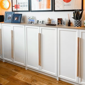 Könnte beinhalten: Weiße Schränke mit Holzgriffen. Die Schränke stehen in einem Raum mit Holzboden. An der Wand über den Schränken hängen drei gerahmte Poster. Die Poster sind alle in Schwarzweiß gehalten und zeigen geometrische Formen. Die Poster sind mit "Bauhaus" und "Führend in der Herrenmode" betitelt.