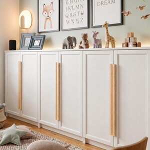 May include: A white storage cabinet with long vertical wooden handles, topped with animal figurines, framed wall art featuring a fox, an alphabet, and the word DREAM, a star-shaped cushion, a knit whale toy, and a wooden chair on a textured rug.