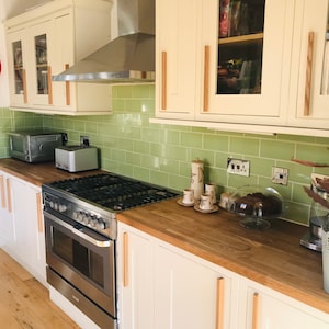 Könnte beinhalten: Helle Küche mit weißen Schränken, Holzarbeitsplatten und einer grünen gefliesten Rückwand. Geräte umfassen einen Edelstahl-Backofen, einen Toaster und einen Toaster-Ofen. Eine rot umrandete Uhr hängt an der Wand. Eine Topfpflanze steht auf der Theke.