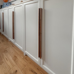 Peut inclure: Armoire blanche avec six portes et poignées en bois. L'armoire est en bois et a une finition blanche. Les portes sont toutes fermées et les poignées sont toutes dans la même position. L'armoire est posée sur un sol en bois.