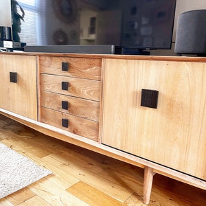 Puede incluir: Un centro de entretenimiento de madera clara con cuatro cajones y dos puertas de armario. Las puertas del armario tienen pomos cuadrados negros. El centro de entretenimiento tiene un acabado de madera clara y se apoya en cuatro patas.