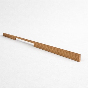 May include: A long, rectangular wooden cabinet pull with a natural wood grain finish. The pull features a handle extending from a wider base, designed for easy gripping. The overall length is approximately 12 inches.