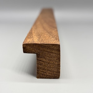 May include: A long, straight piece of dark brown wood with a distinct L-shaped profile. The wood features a visible, natural grain pattern throughout its surface. The end of the piece shows a sharp, rectangular cross-section with a protruding edge.