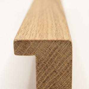 May include: A long, linear cabinet pull made of light brown oak wood with a visible natural grain pattern. The handle features an L-shaped profile with a flat top surface and a vertical front edge, designed for mounting onto a flat cabinet door surface.