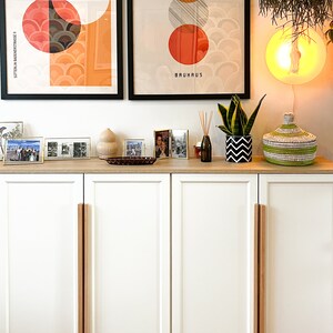 以下が含まれることがあります： 木製の取っ手が付いた白い扉が2つあるキャビネット。キャビネットは明るい木製の天板があり、額縁に入った写真、植物、装飾品で飾られています。