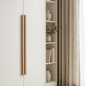 May include: A tall white wardrobe featuring long, vertical light wood handles. Beside the wardrobe is a built-in shelving unit containing books, a small wooden bird figurine, ceramic vases, and a green plant. A beige knit blanket sits nearby.