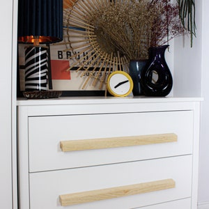 Könnte beinhalten: Weiße Kommode mit drei Schubladen, jede mit einem hellen Holzgriff. Auf der Oberseite befinden sich eine marineblaue Lampe, ein dekorativer Spiegel, eine gelbe Uhr und Vasen mit getrockneten Blumen. Ein Bauhaus-Ausstellungsplakat ist sichtbar.