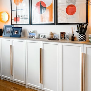 May include: White cabinet with four doors and wooden handles. The cabinet is against a white wall with three framed posters featuring geometric shapes and the text "FÜHREND IN DER HERRENMODE", "BAUHAUS EXHIBITION JULY 23, 19:1", and "BAUHAUS".